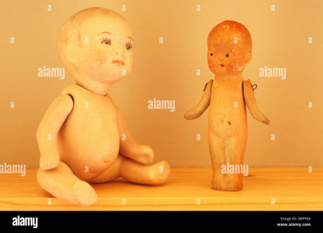 Close Up Of Two Naked Traditional Baby Dolls From 1920s On Wooden Shelf With One Sitting And Other Standing Looking At Her Photo
